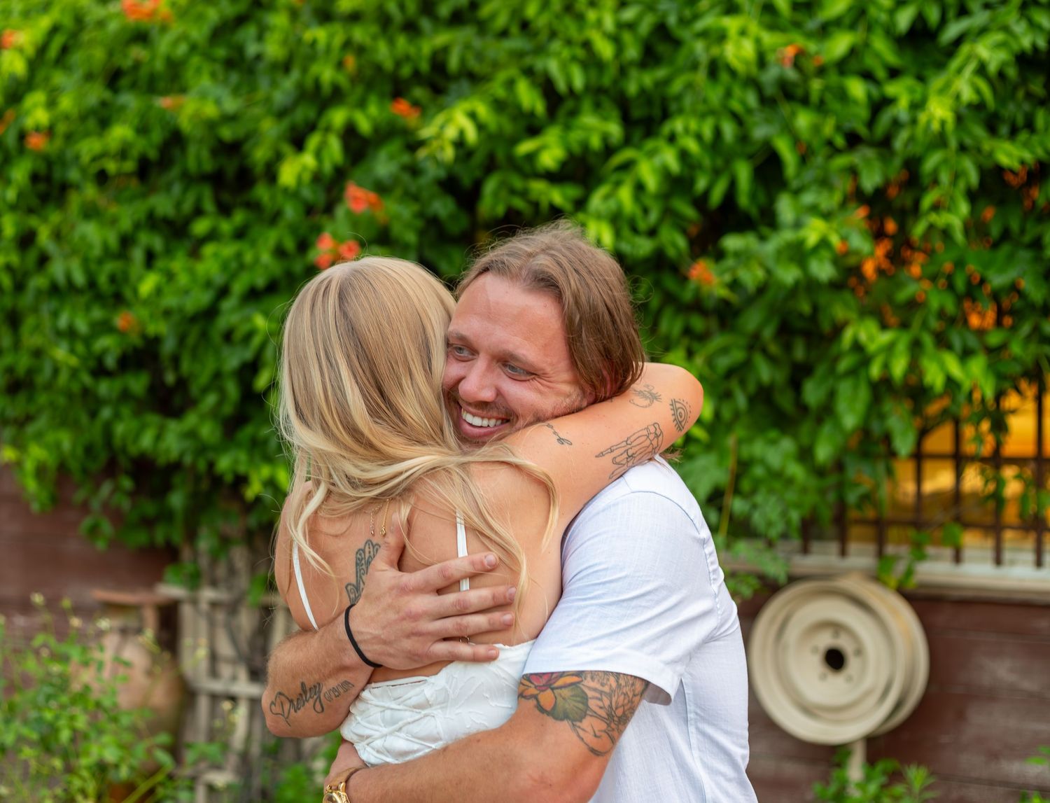Tattooed man smiling while hugging a blonde woman outdoors with lush green foliage in the background.