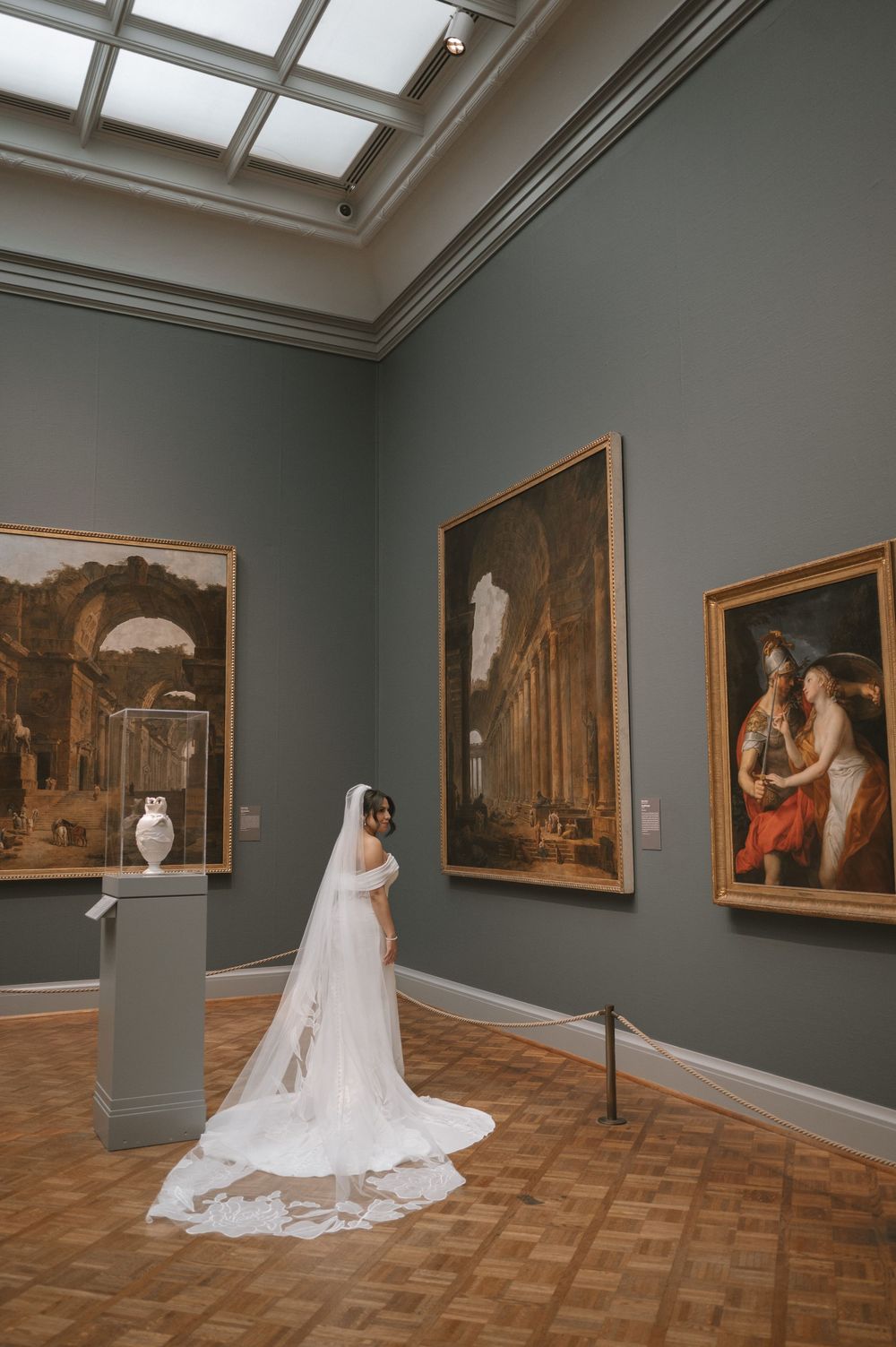Bride in white gown and long veil poses in elegant art museum gallery with classical paintings.