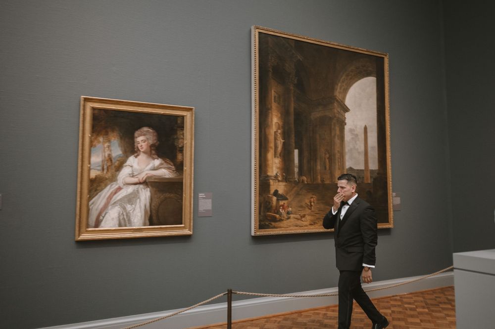 Man in tuxedo walking past classic oil paintings in an elegant museum gallery with gray walls.
