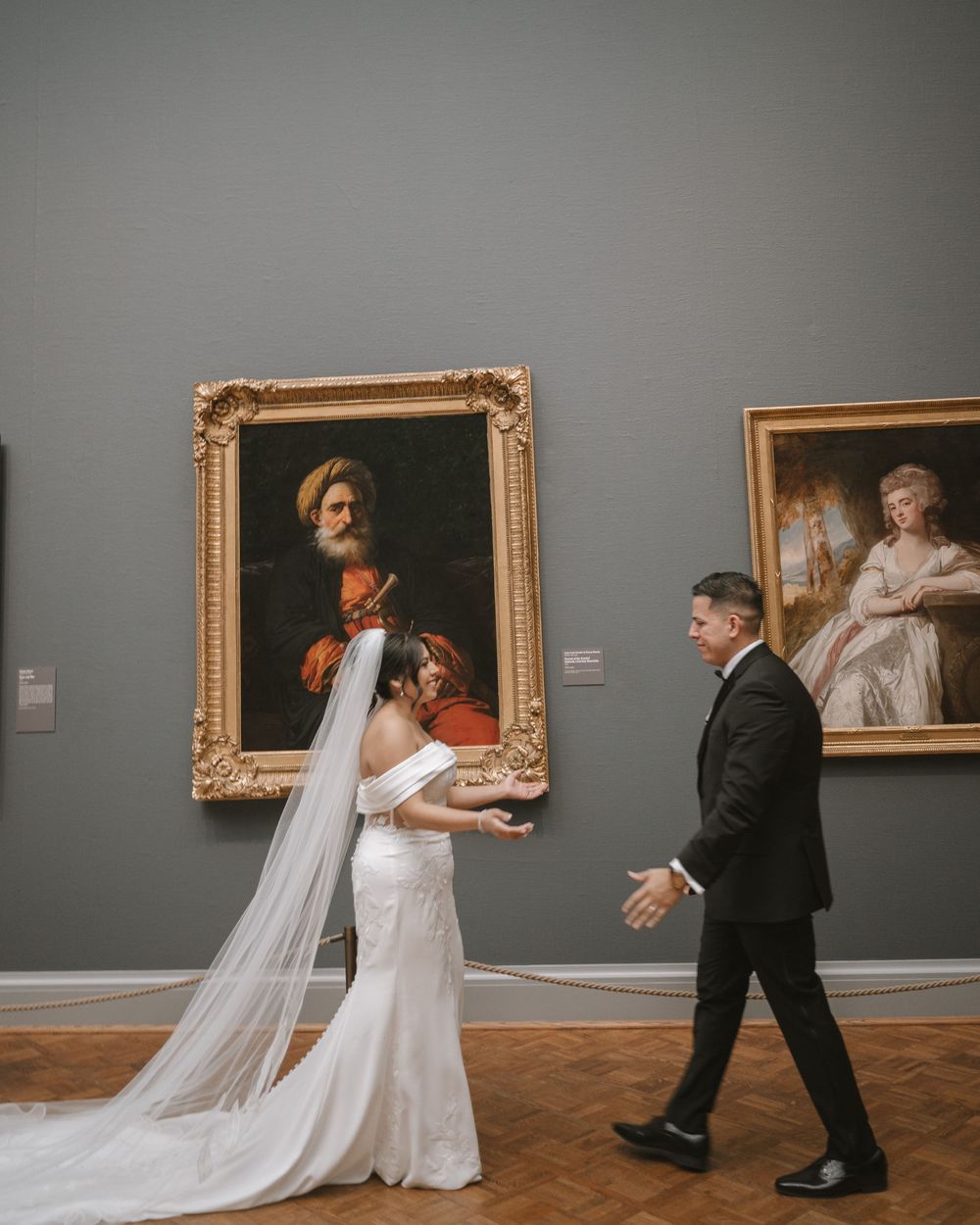 Bride and groom walking through an art museum gallery with classical paintings on gray walls.