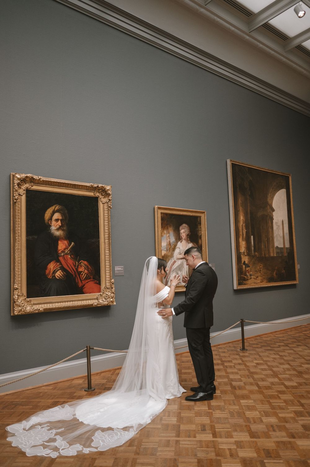 Bride and groom holding hands in an art museum gallery with classical paintings on gray walls.