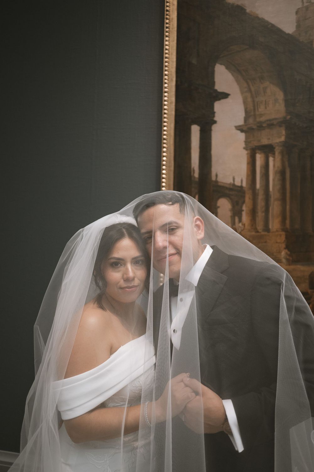 Bride and groom smiling together under a flowing white veil in front of a classical painting.