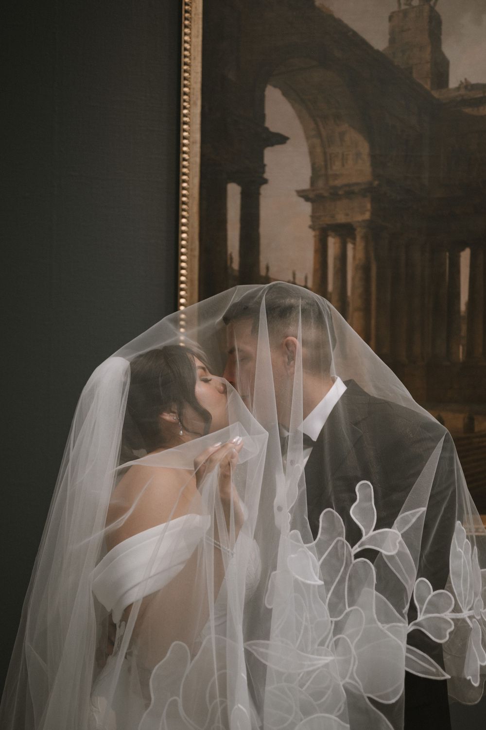 Bride and groom sharing a romantic kiss under an elegant floral veil in front of a classical painting.