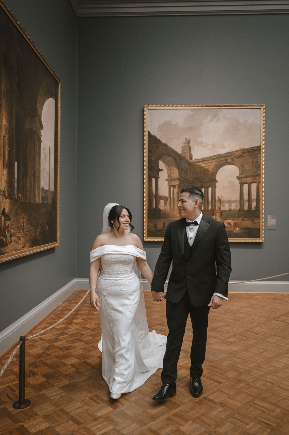 Bride in off-shoulder gown and groom in tuxedo walking hand in hand through an art museum.