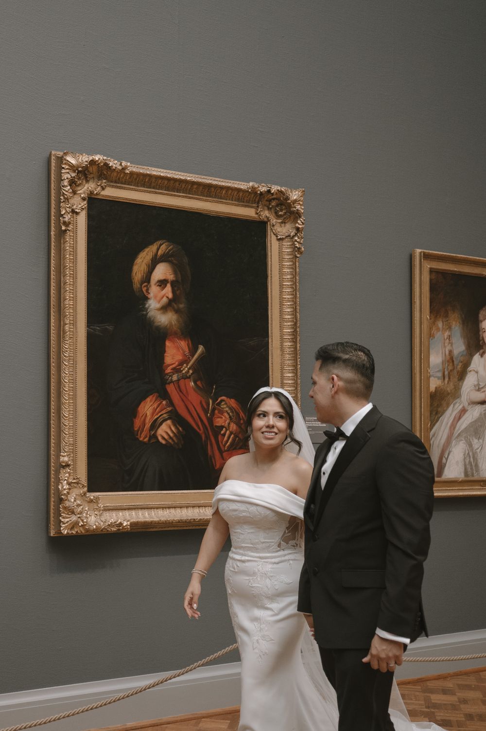 Smiling bride in white gown and groom in tuxedo standing beside a classical portrait painting.
