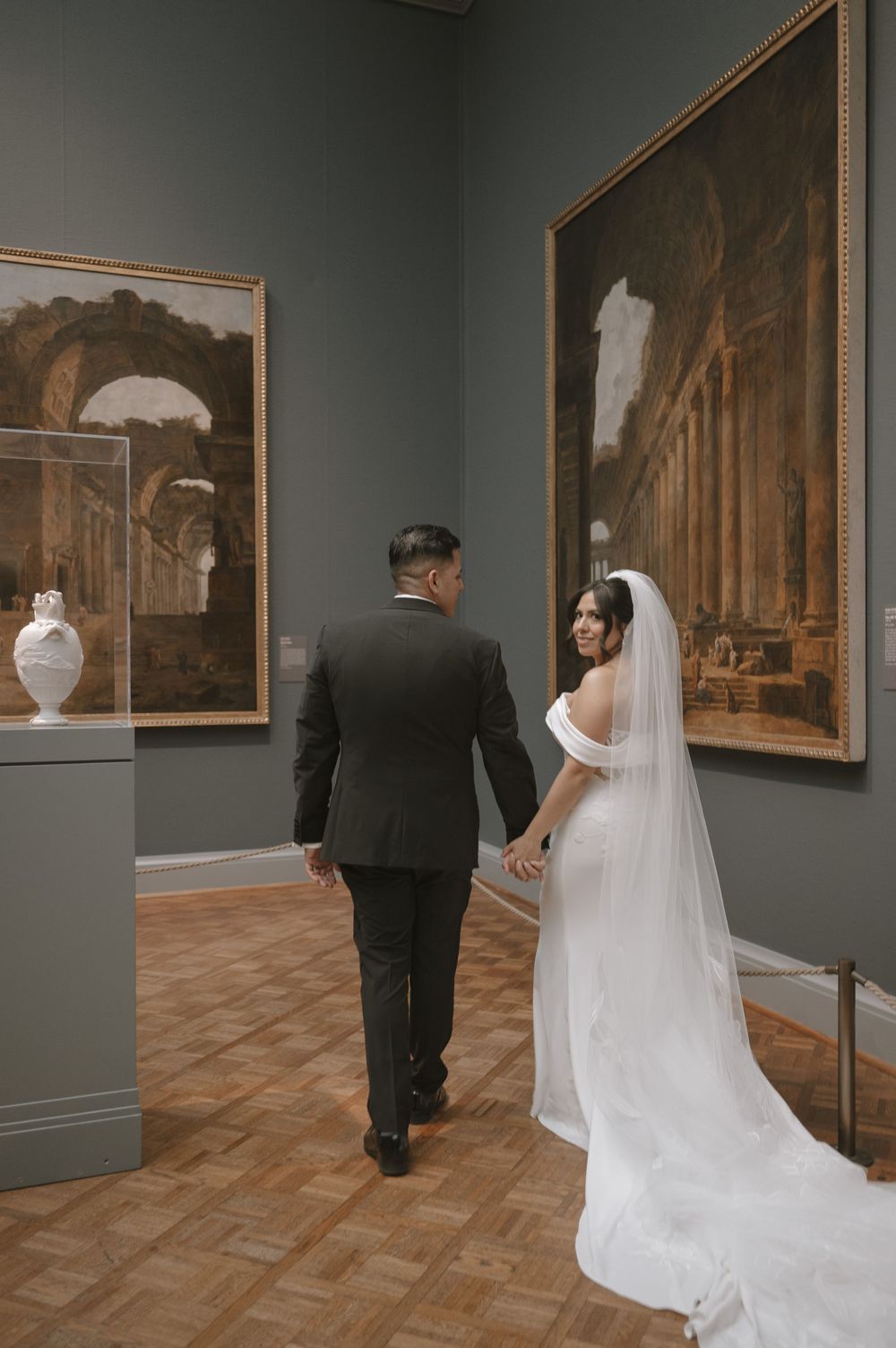 Bride and groom walk hand-in-hand through an art museum gallery, with large classical paintings on gray walls.