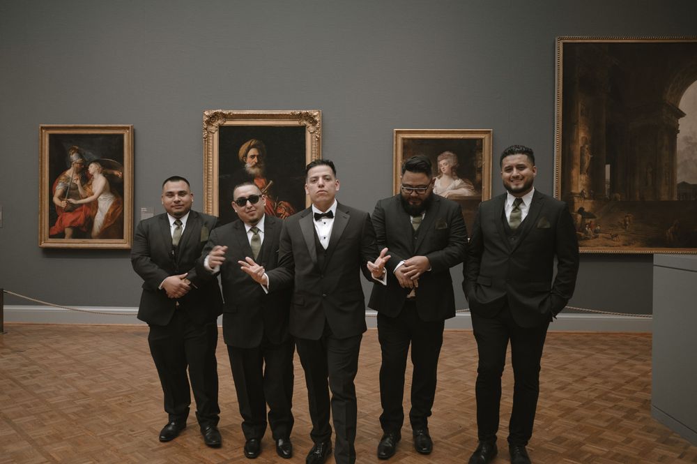 Five men in black tuxedos posing in an art museum gallery with classic paintings on the wall.