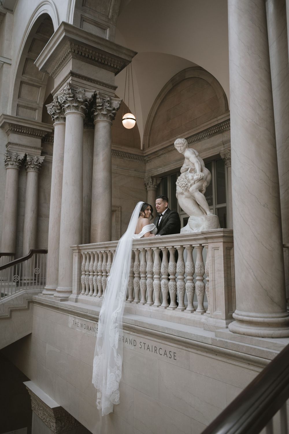 Bride and groom pose on grand marble staircase balcony with classical columns and sculpture at elegant wedding venue.