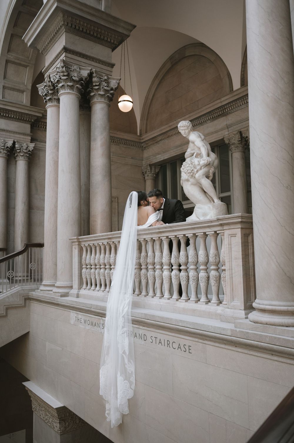 Bride and groom sharing a kiss on a grand marble staircase with classical columns and sculpture.