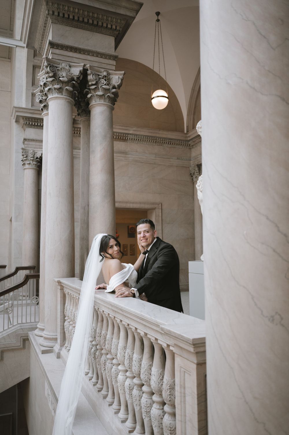 Bride and groom posing on marble balustrade with grand columns in a neoclassical building.