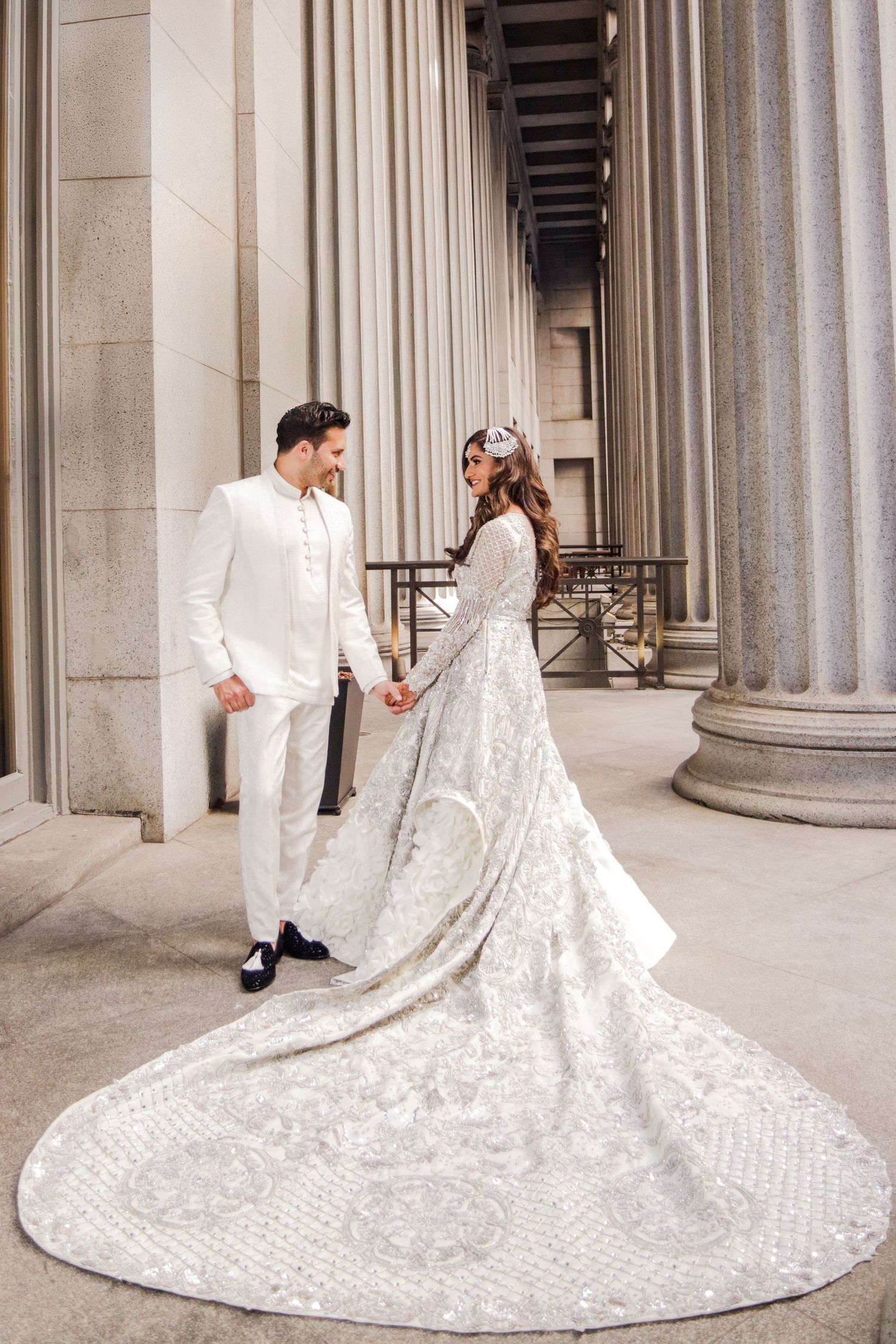 Elegant couple in white wedding attire holding hands among grand columns, bride wearing a stunning embroidered gown with long train.