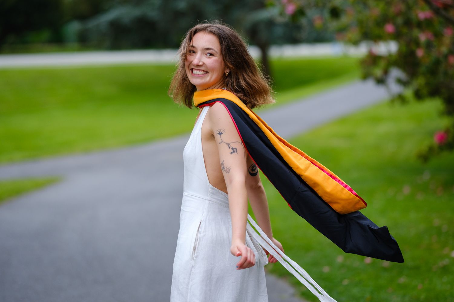 Smiling graduate in white dress with black and gold academic hood poses outdoors on a tree-lined path.
