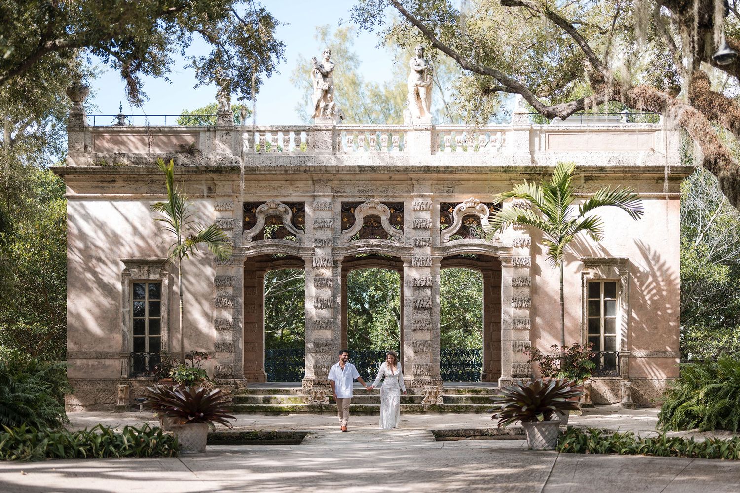 Bride and groom walk hand in hand through Vizcaya Gardens, framed by historic architecture, tropical plants, and elegant stone details.