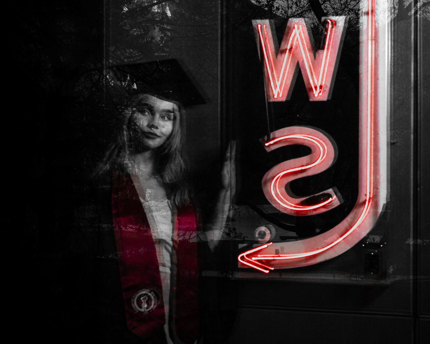 Black and white photo of a graduate in cap and red sash posing near a glowing neon WS sign.