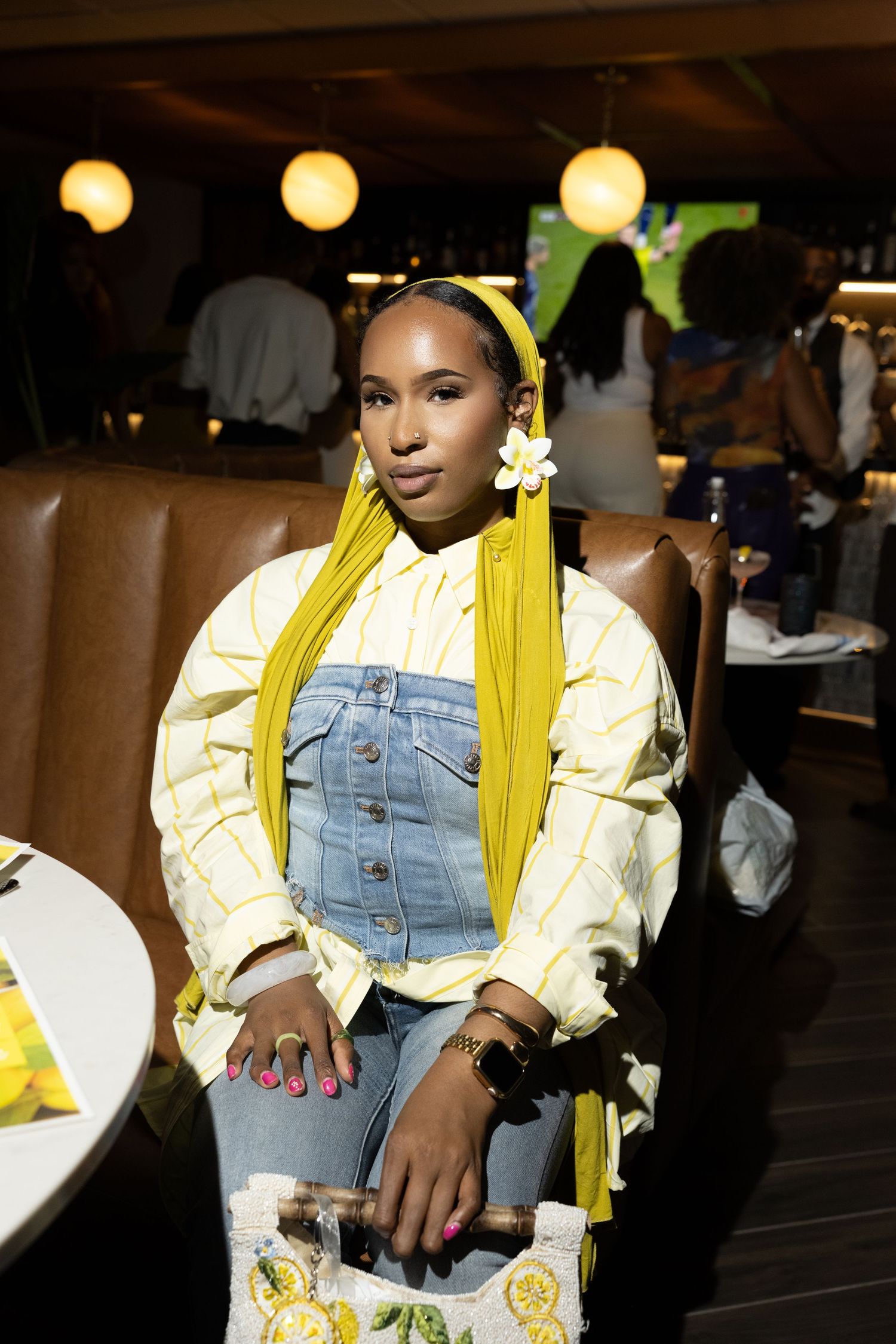 Young woman in yellow headscarf, denim overalls, and striped jacket seated in a stylish restaurant setting.