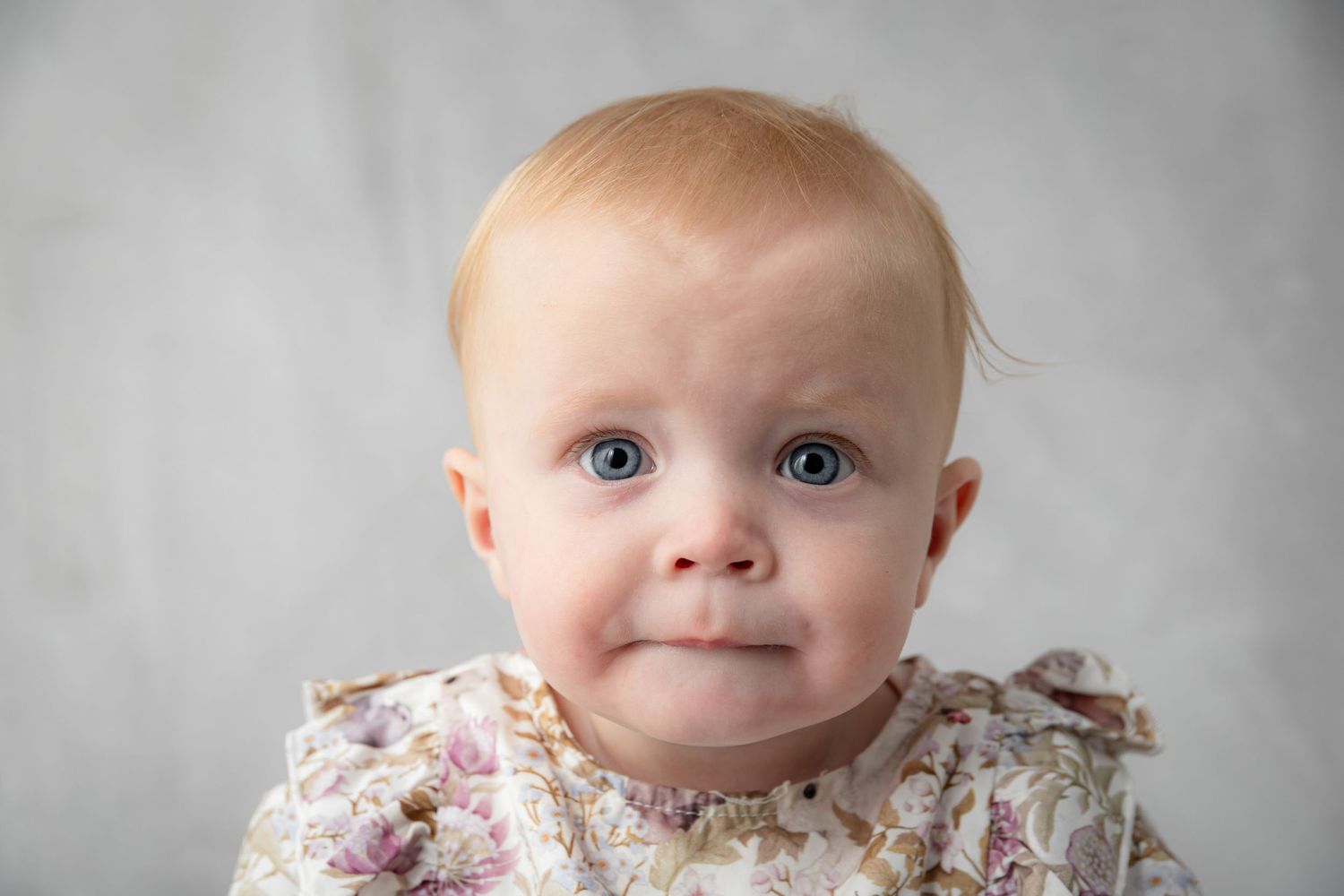 Adorable baby girl with blue eyes and red hair wearing a floral dress, posed against a gray background.