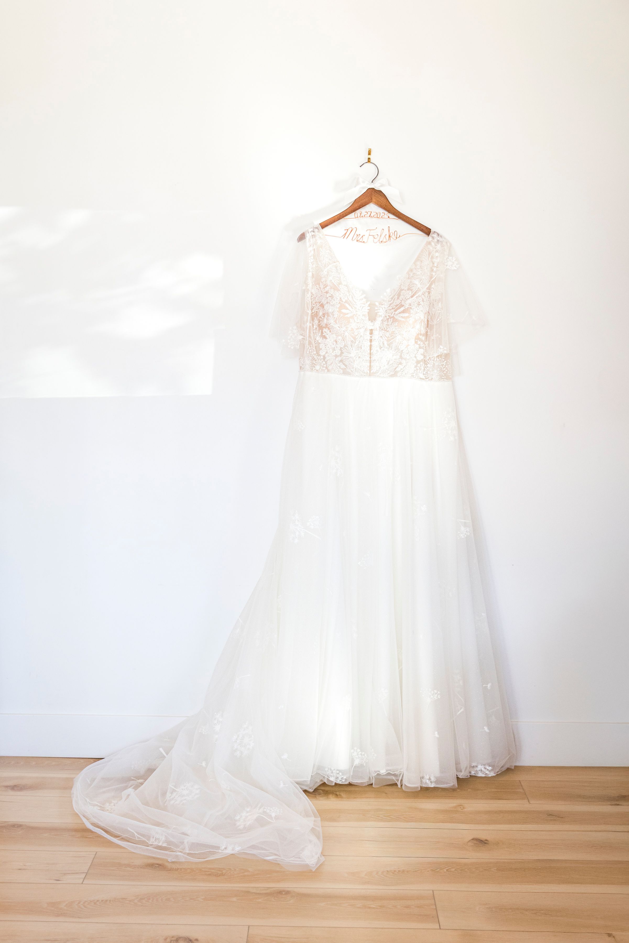 Elegant white lace wedding dress with flutter sleeves and flowing tulle skirt on wooden hanger against white wall.