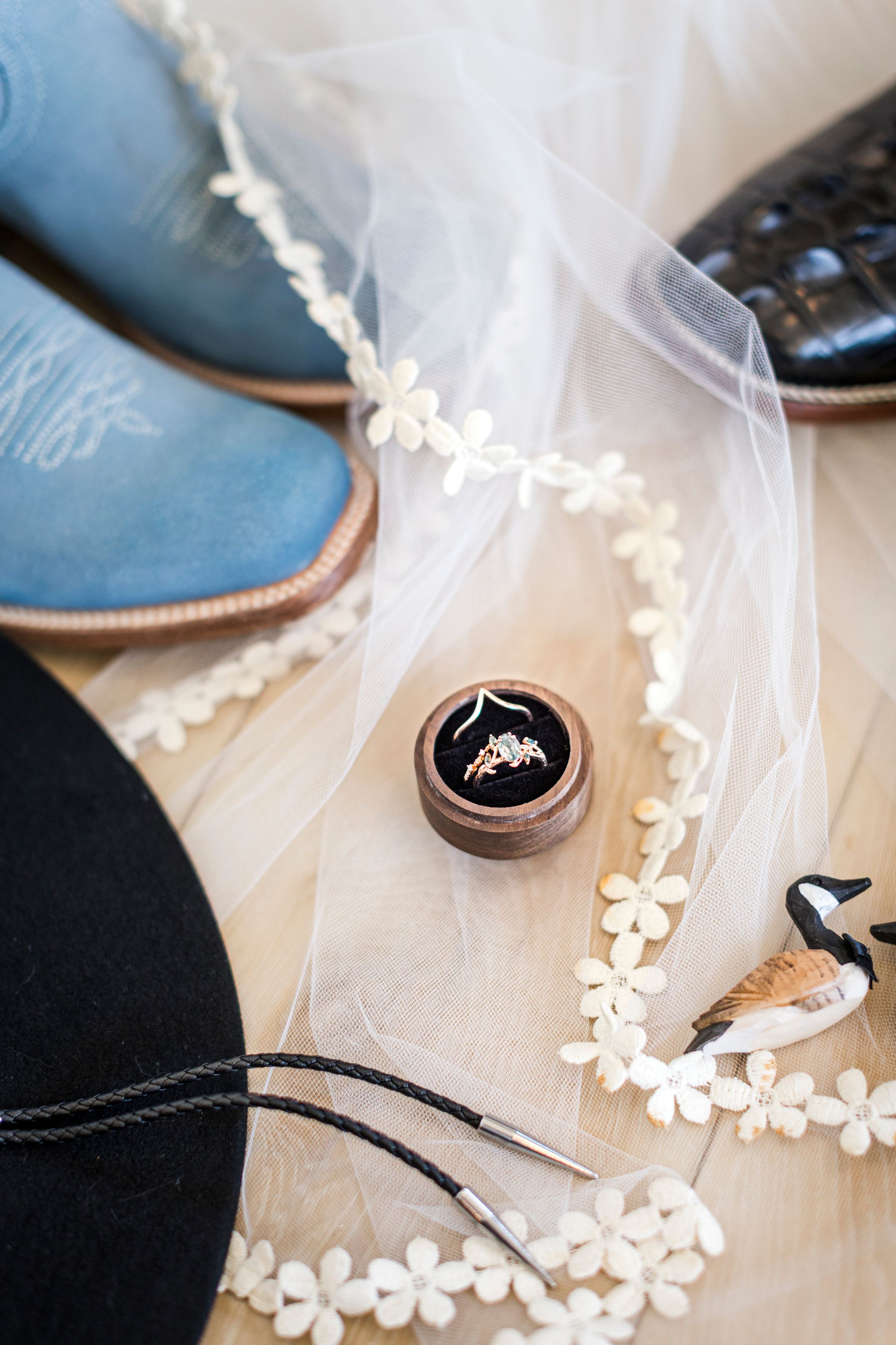 Wedding flat lay with blue cowboy boots, lace veil, ring box, bolo tie, and black hat on wood floor.