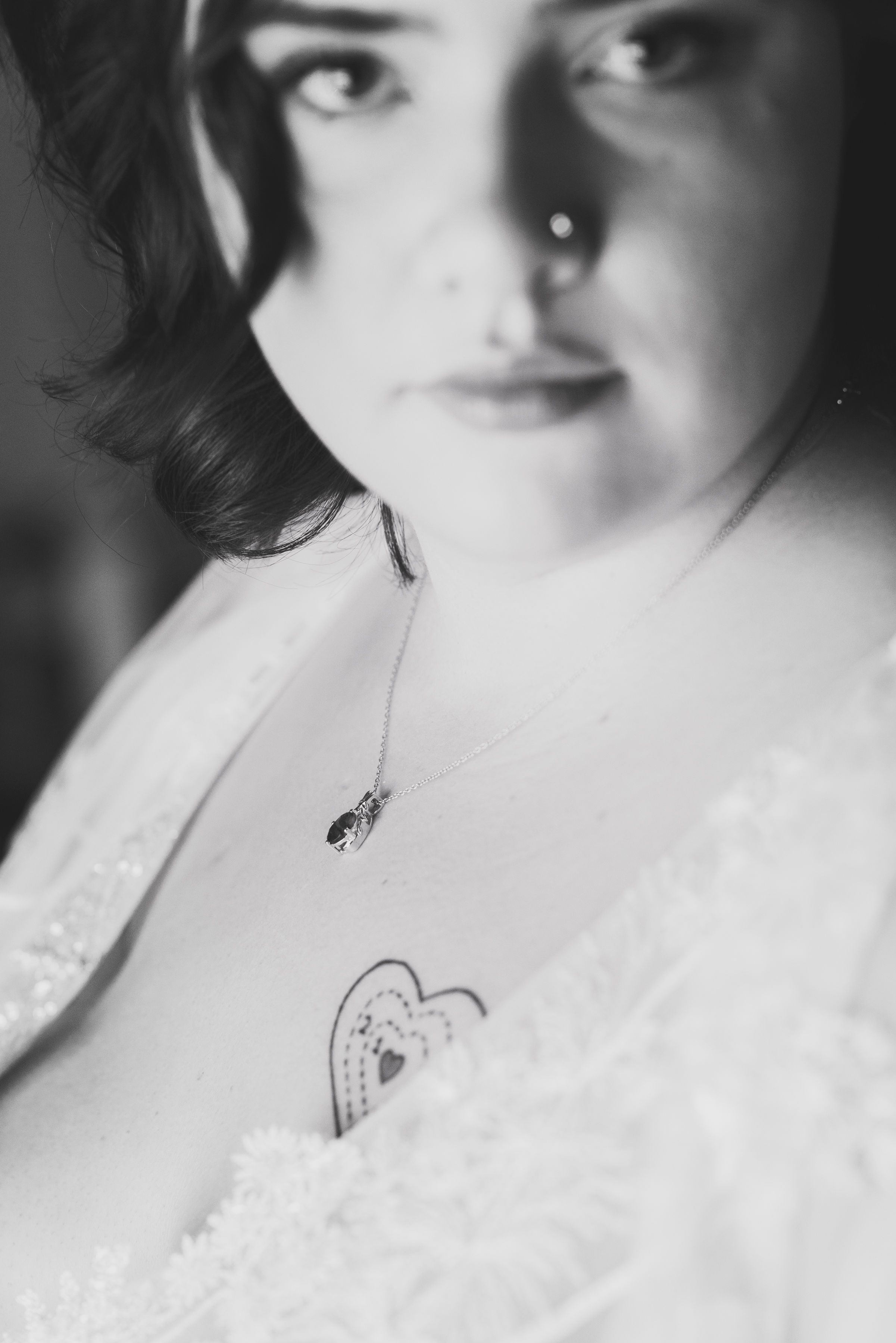 Black and white portrait of a woman with a heart tattoo on her chest wearing a delicate necklace and white lace top.