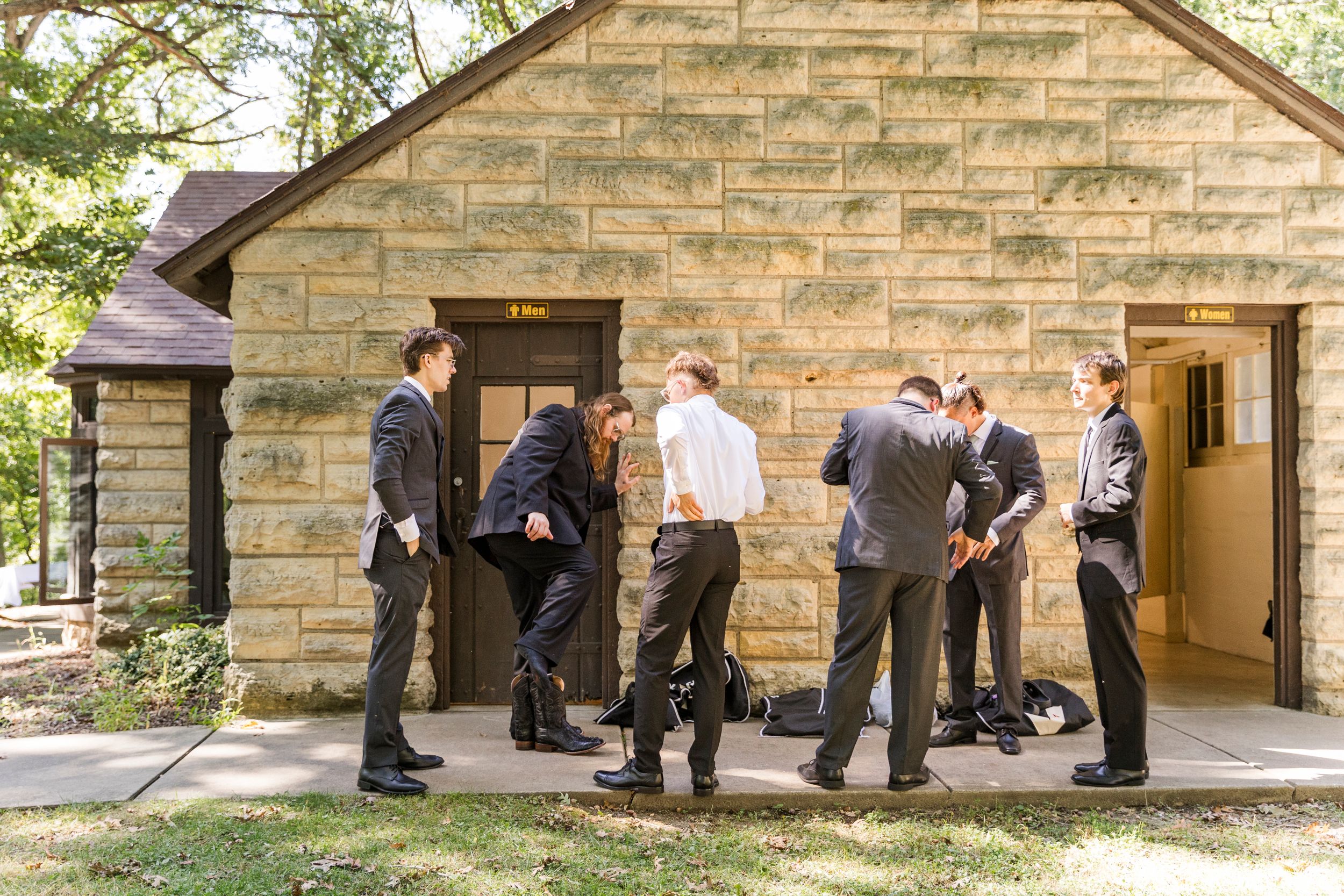 Groomsmen in suits gather outside a stone building before a wedding ceremony.