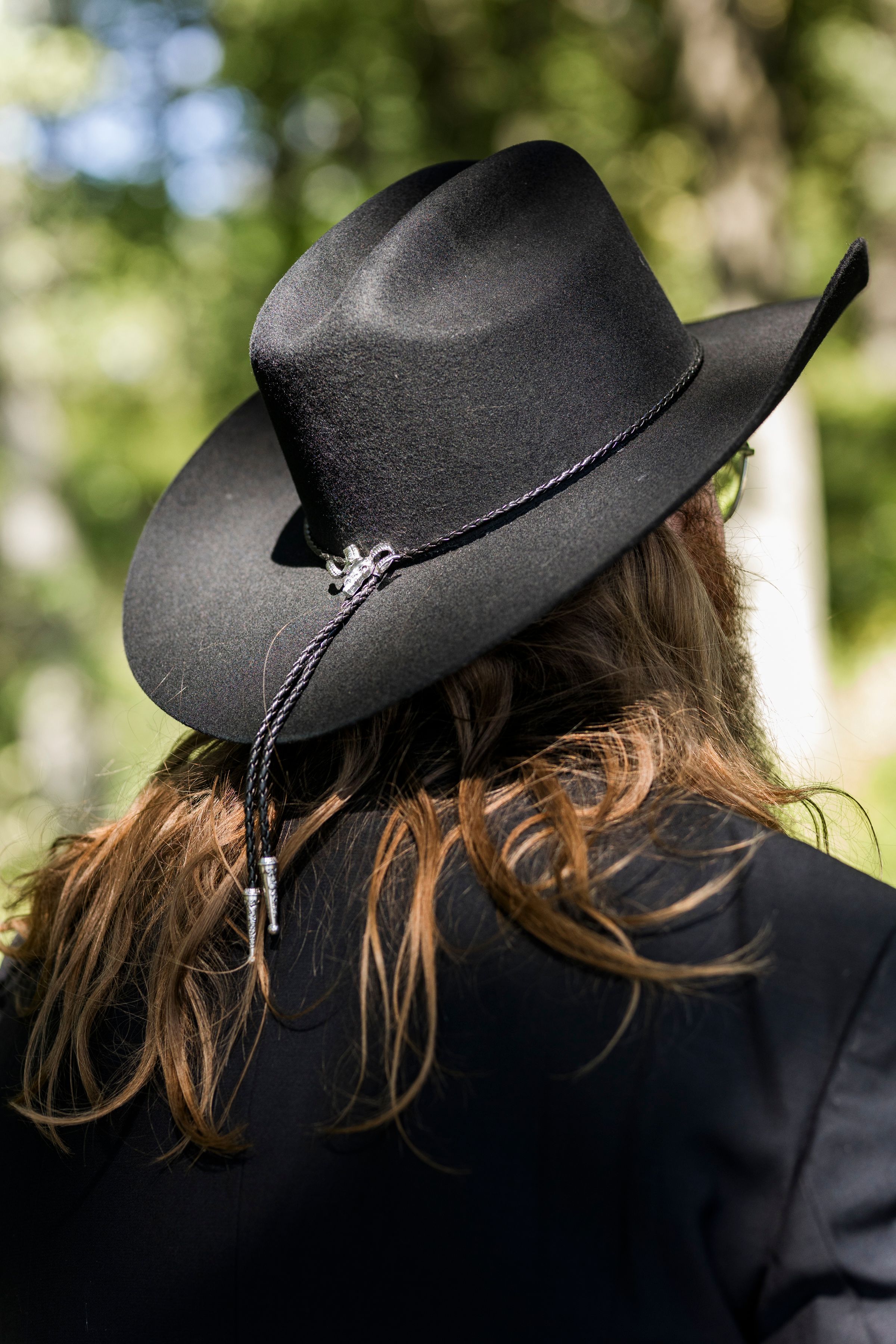 Back view of a person with long brown hair wearing a black cowboy hat with decorative bolo tie band outdoors.