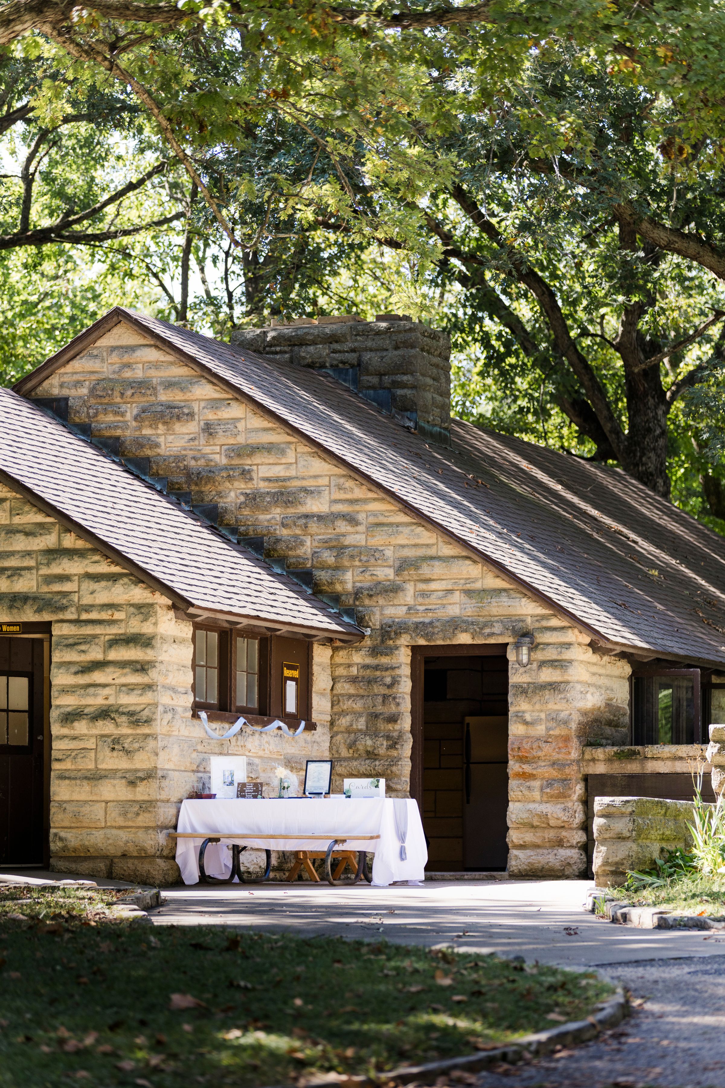 Rustic stone cottage with shingled roof, chimney, and outdoor reception table set under large shade trees.