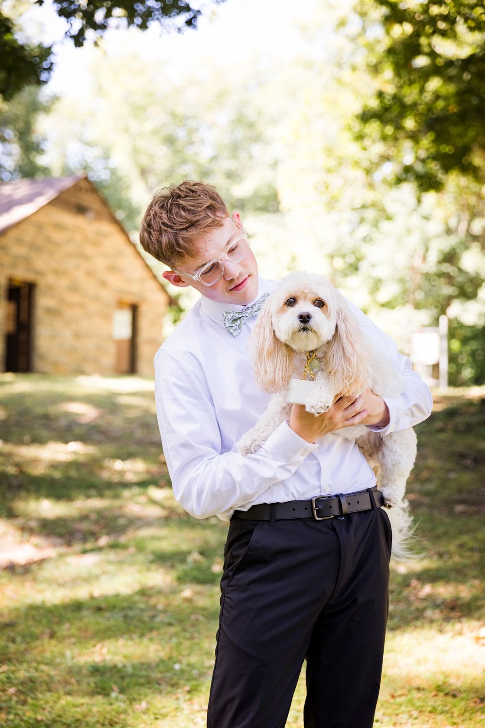 Young man in white shirt and bow tie holding a fluffy white dog outdoors at a wedding venue.