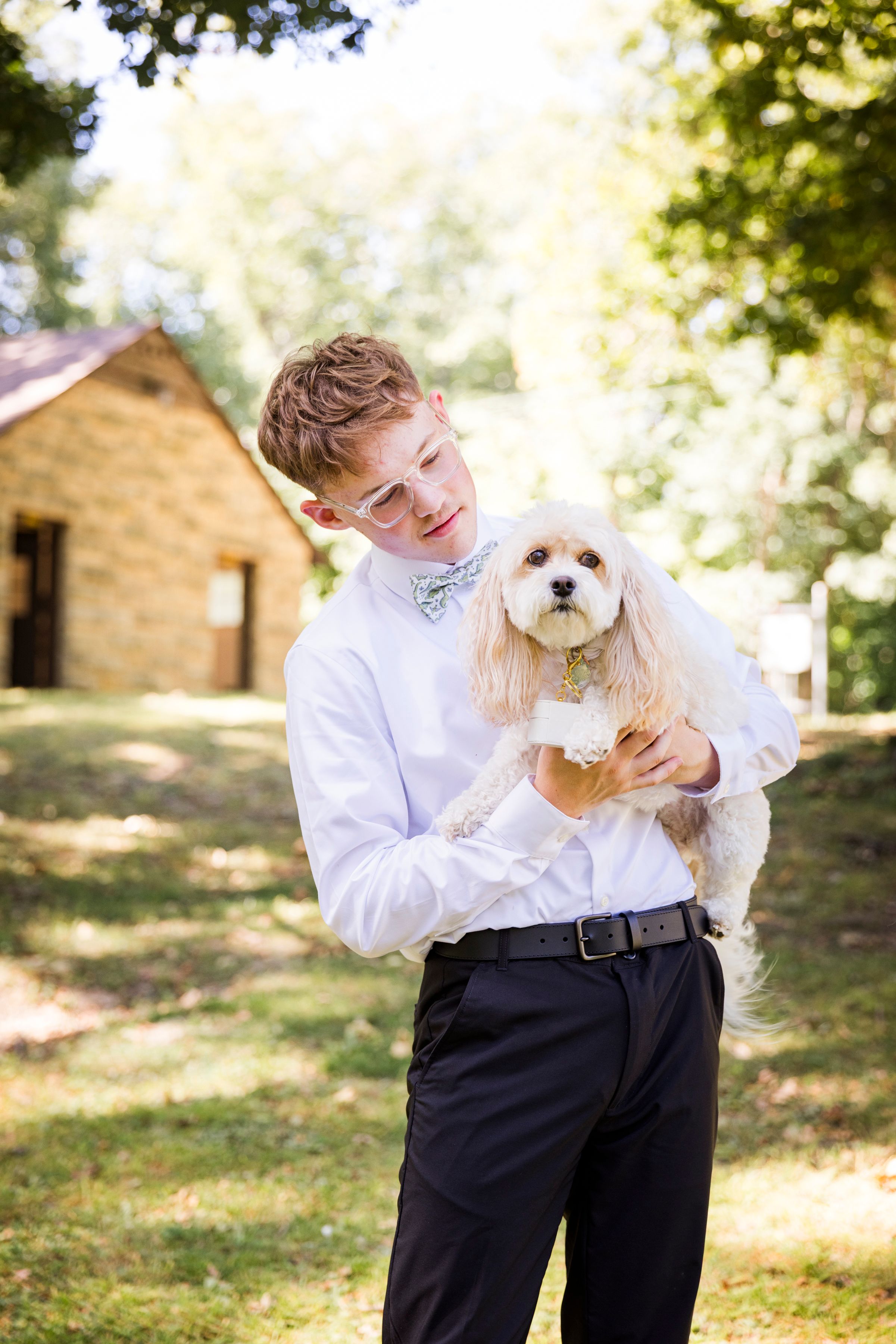 Young man in white shirt and bow tie holding a fluffy white dog outdoors at a wedding venue.