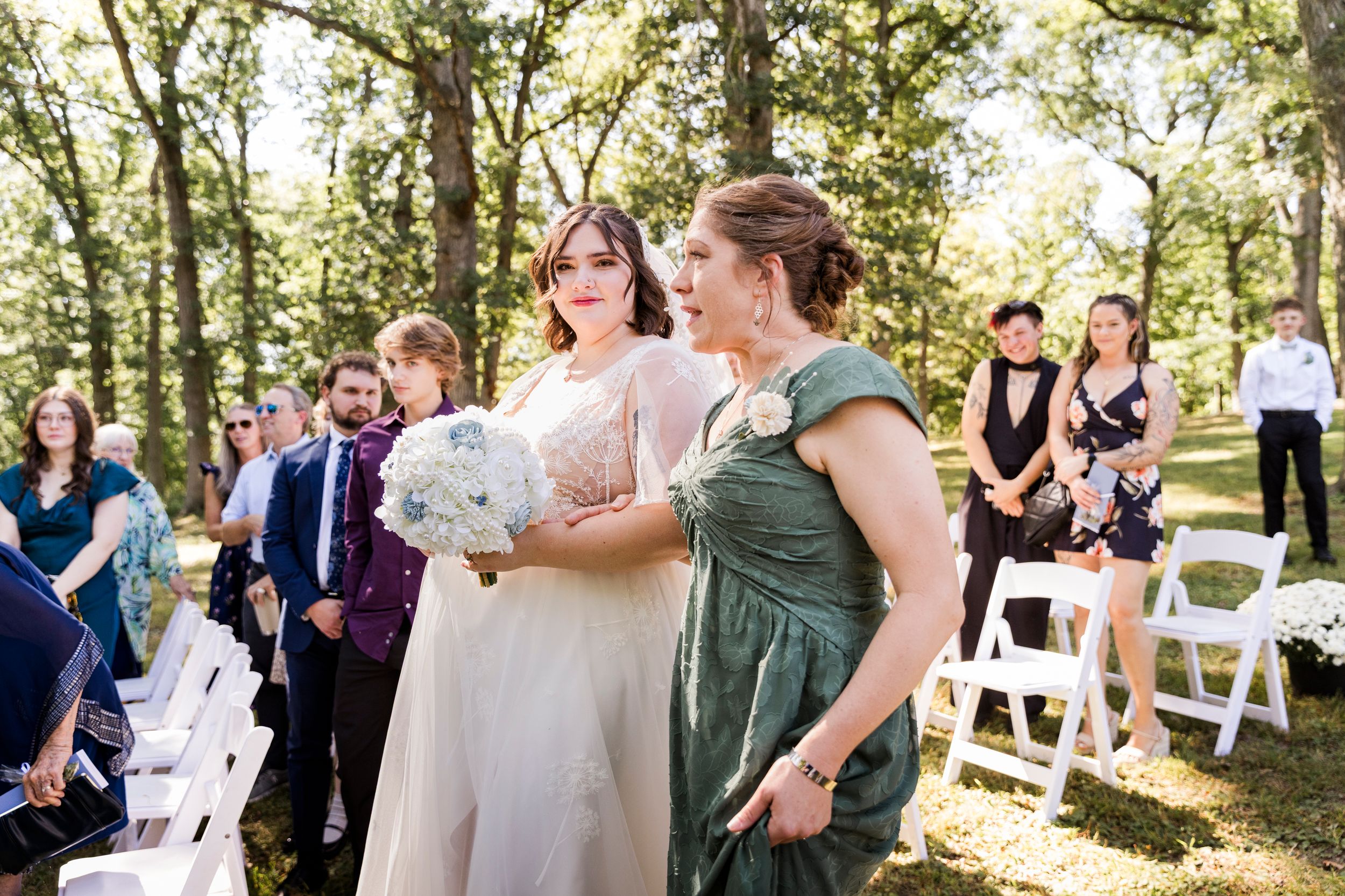 Bride in blush gown walks down outdoor aisle with bridesmaid in sage green dress, holding white bouquet.