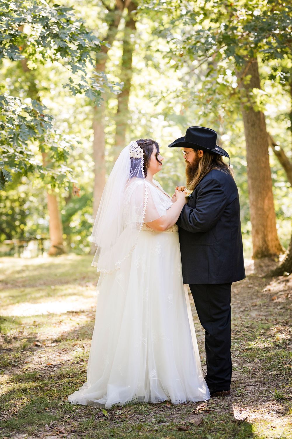 Bride in white gown and veil faces groom in black suit and cowboy hat in a sunlit forest setting.