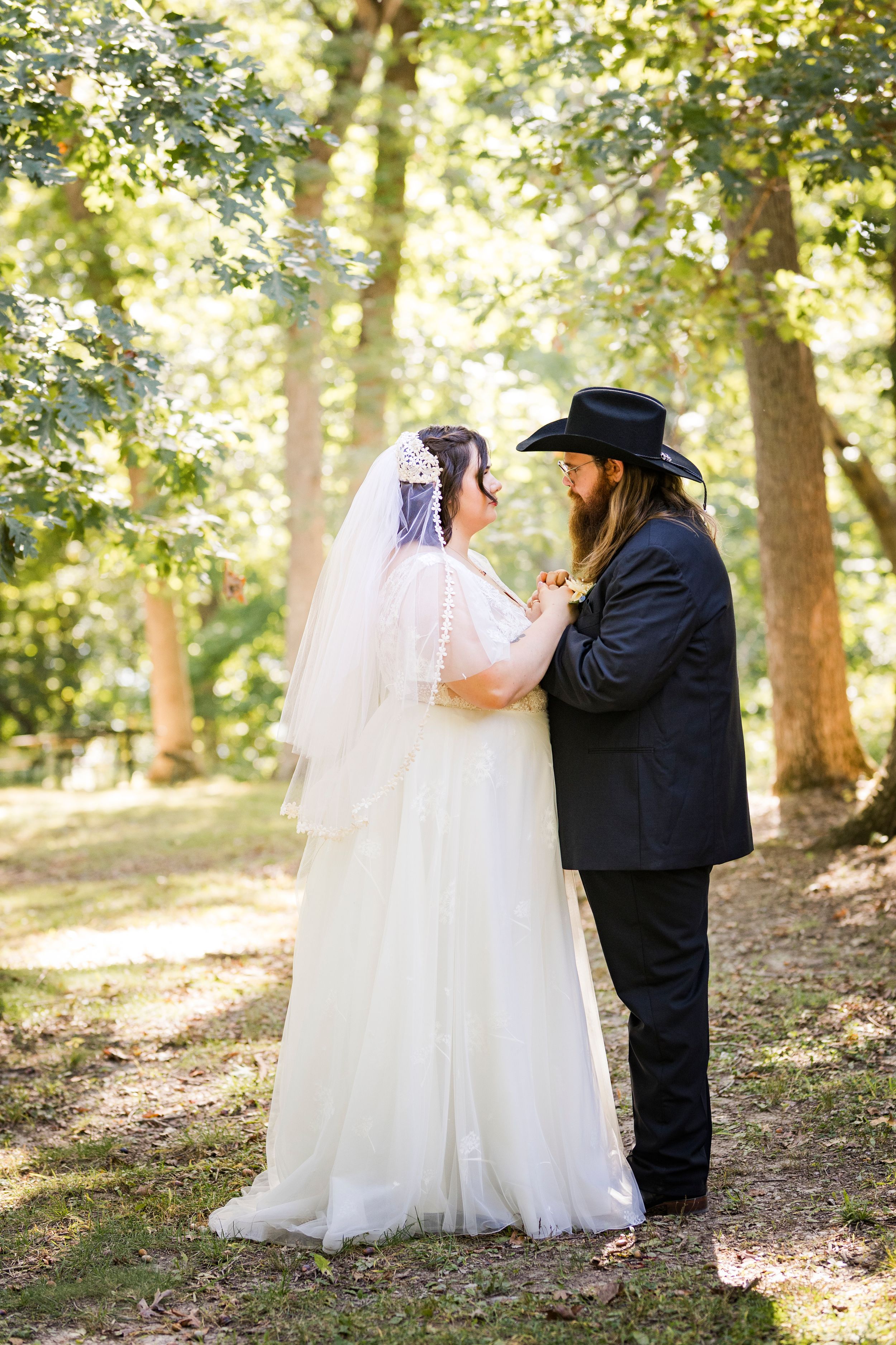 Bride in white gown and veil faces groom in black suit and cowboy hat in a sunlit forest setting.