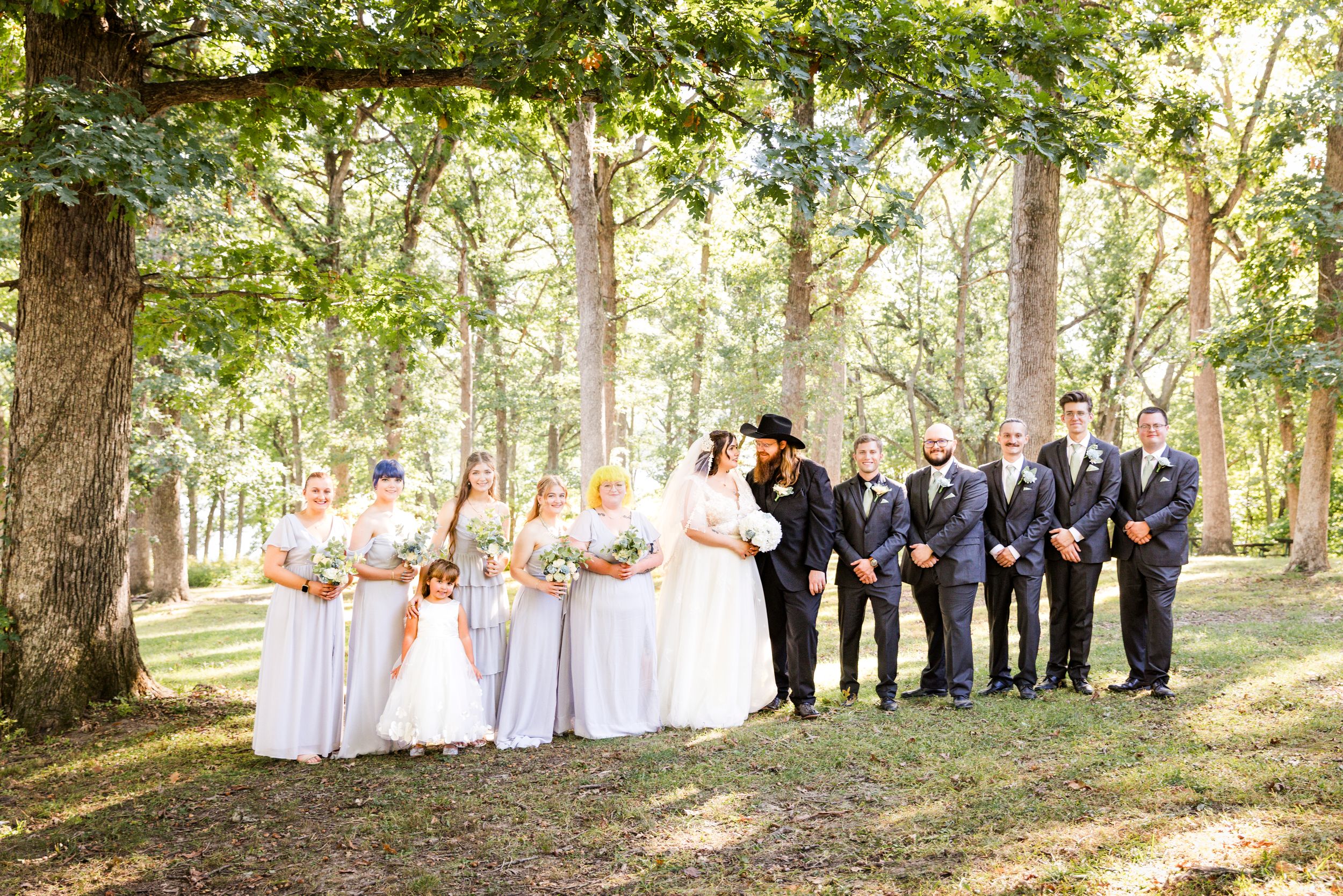 Wedding party poses outdoors among tall trees, bridesmaids in gray dresses and groomsmen in navy suits flanking the bride and groom.