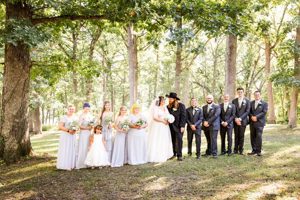 Wedding party poses outdoors among tall trees, bridesmaids in gray dresses and groomsmen in navy suits flanking the bride and groom.