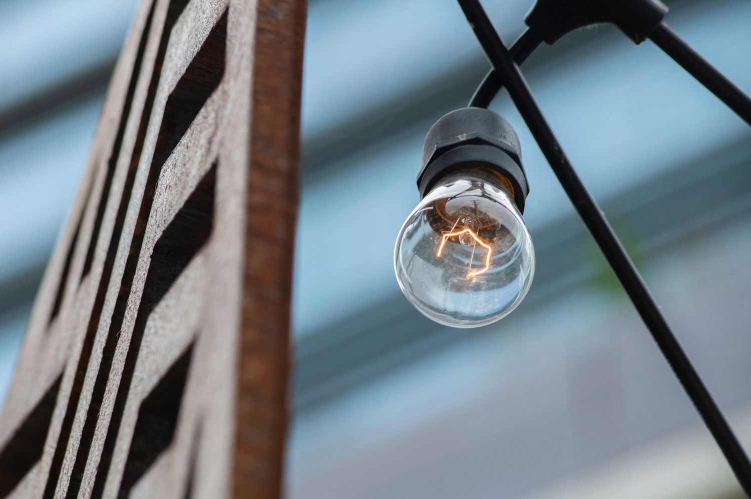 Vintage Edison bulb hanging on string lights outdoors with wooden post and glass ceiling backdrop.