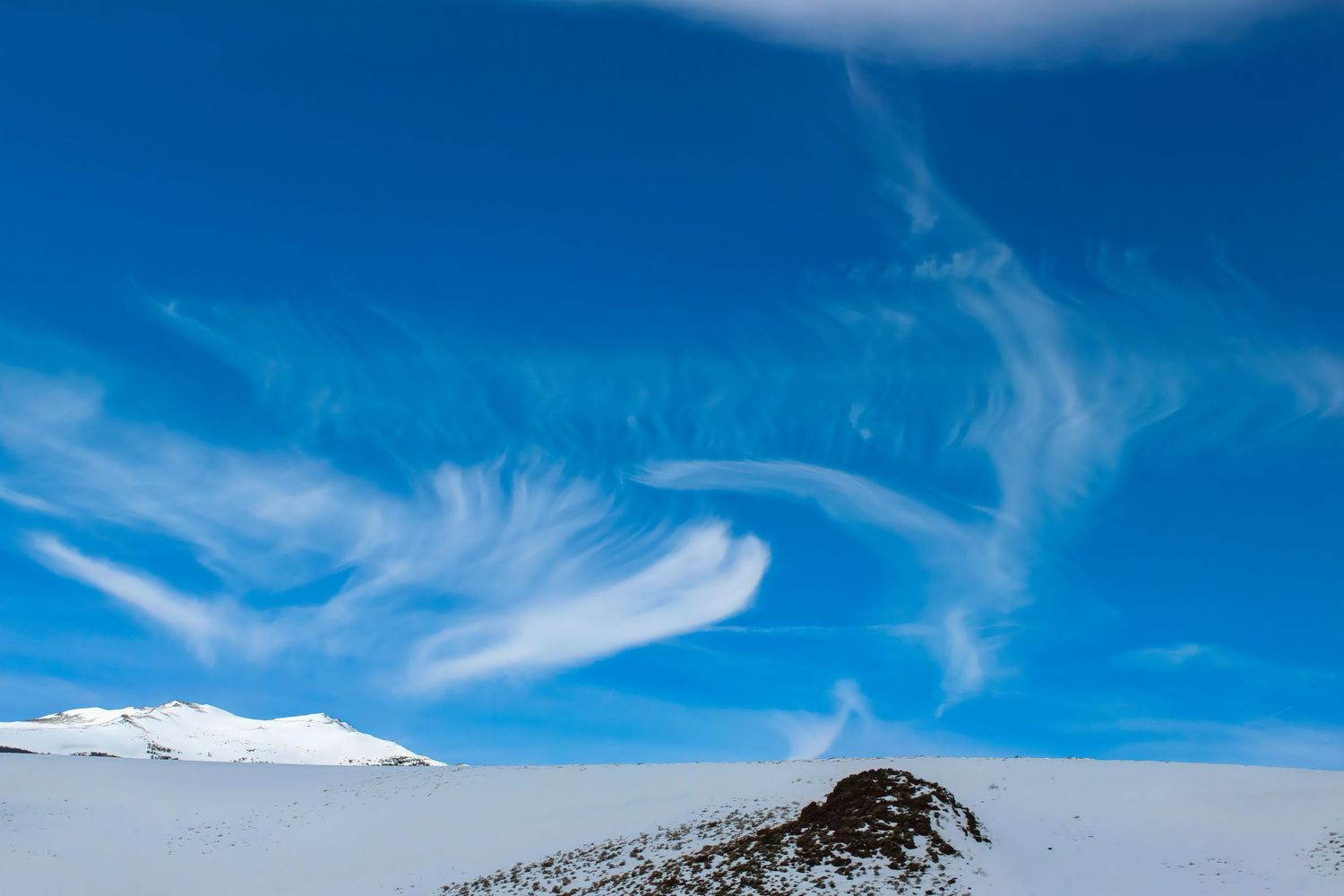 Snow-covered mountain landscape with dramatic wispy cirrus clouds swirling across a vivid blue sky.
