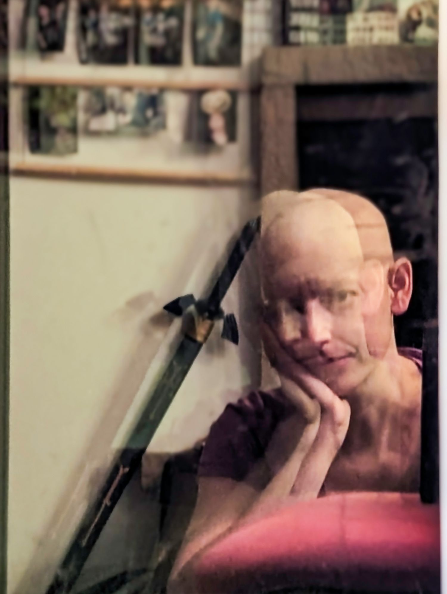 Bald man resting chin on hand, seated in a pink chair, with photos displayed on a shelf in the background.