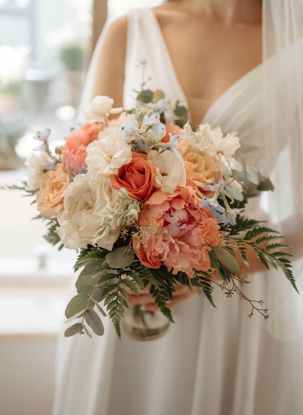 Bride holding a lush wedding bouquet with peach roses, white peonies, and greenery in a soft, romantic arrangement.