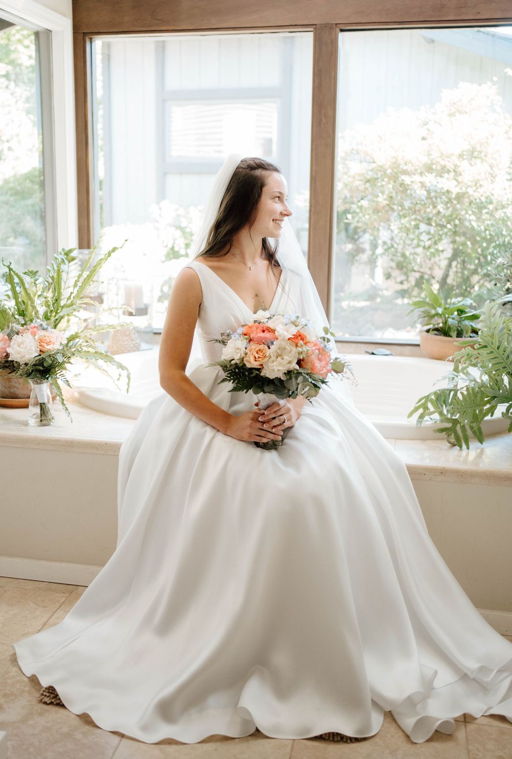 Bride in white ball gown holding peach and white bouquet, seated by sunlit window with greenery.