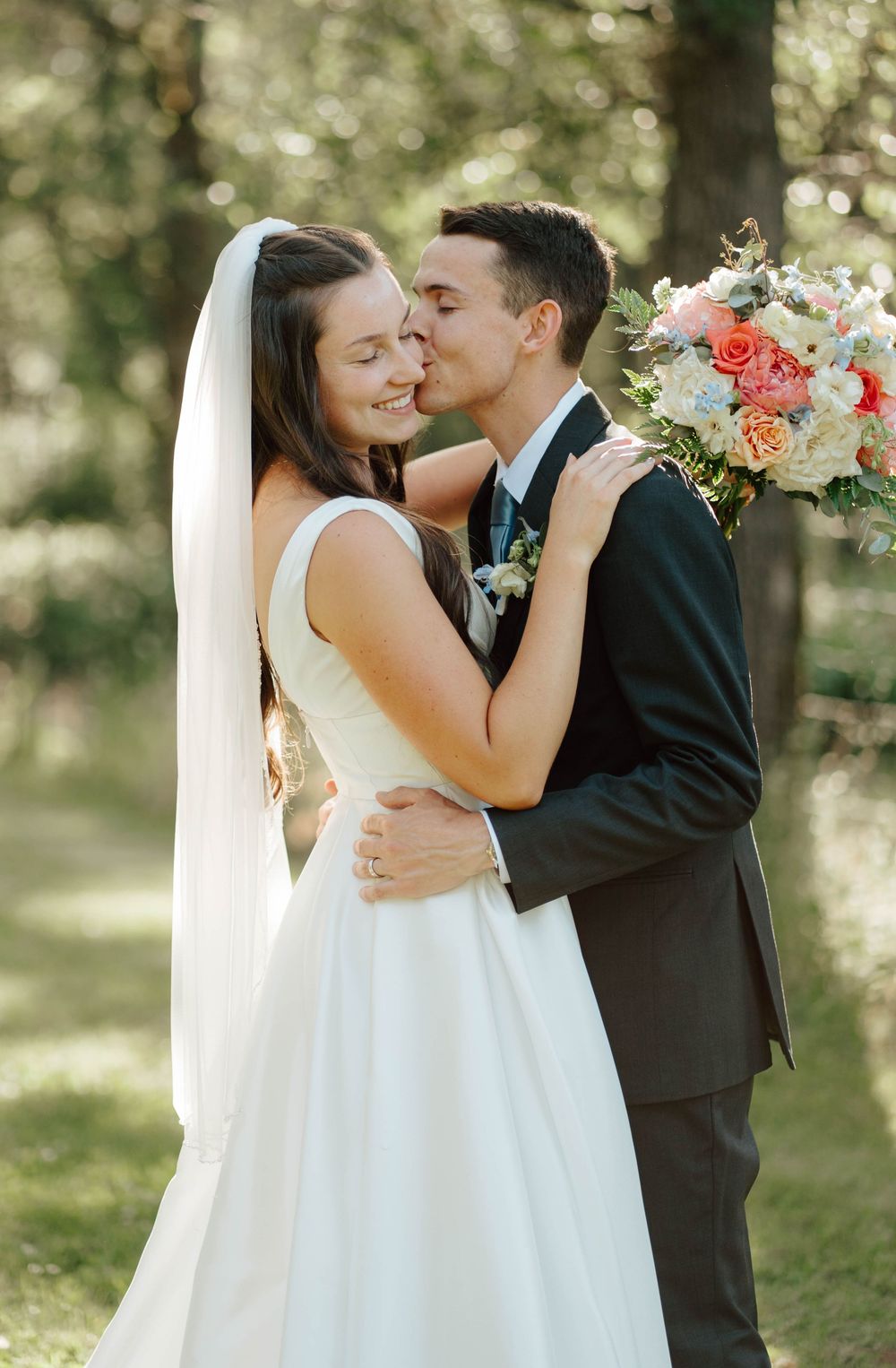 Groom tenderly kisses bride on cheek as she smiles holding a pink and white bouquet in a sunlit garden.