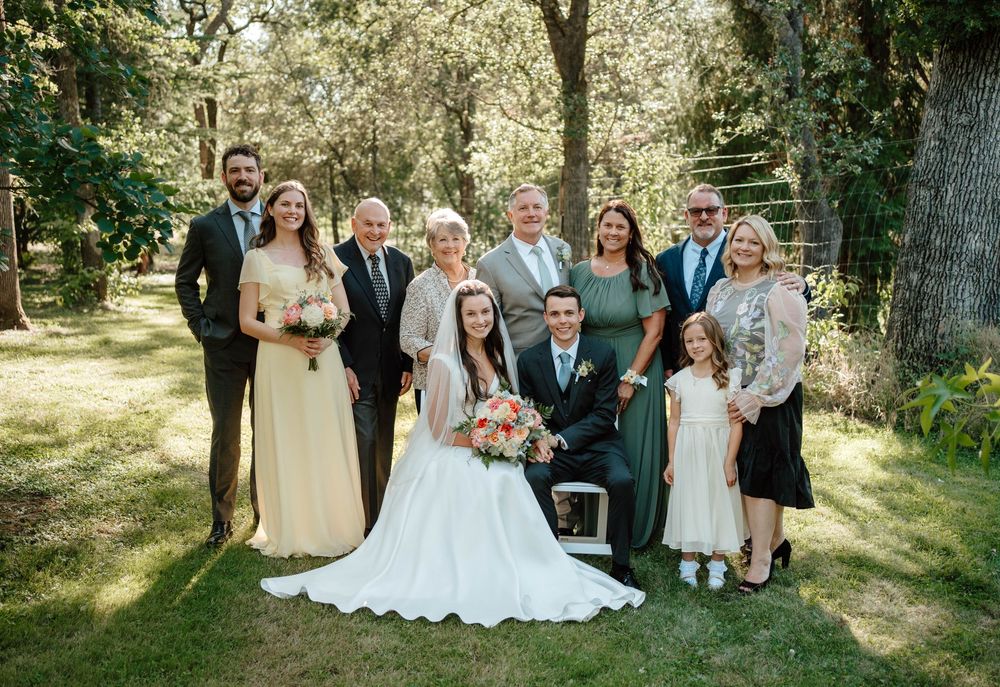 Wedding party posing outdoors on green lawn with bride and groom centered, surrounded by family in formal attire.