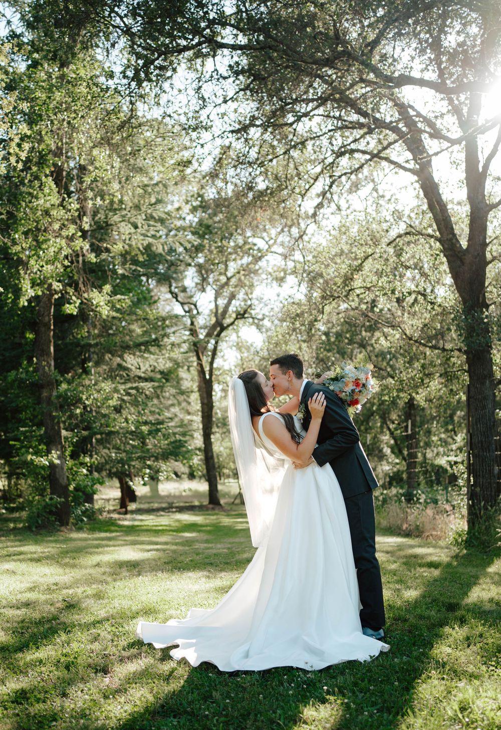 Bride and groom share a romantic kiss in a sunlit forest clearing, bride in white gown with long train holding bouquet.