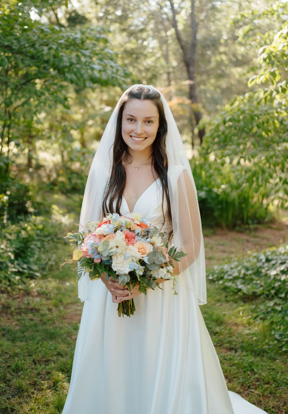 Smiling bride in white gown and veil holds colorful bouquet of peach and white flowers outdoors in lush green garden.