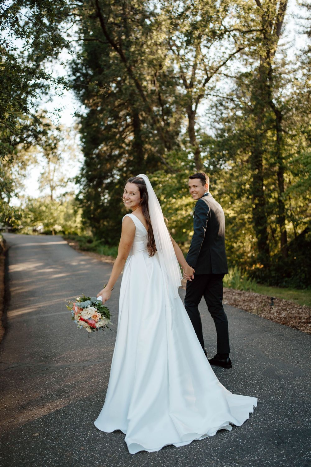 Bride and groom holding hands on a tree-lined road, bride in white gown with veil holding bouquet, groom in dark suit.