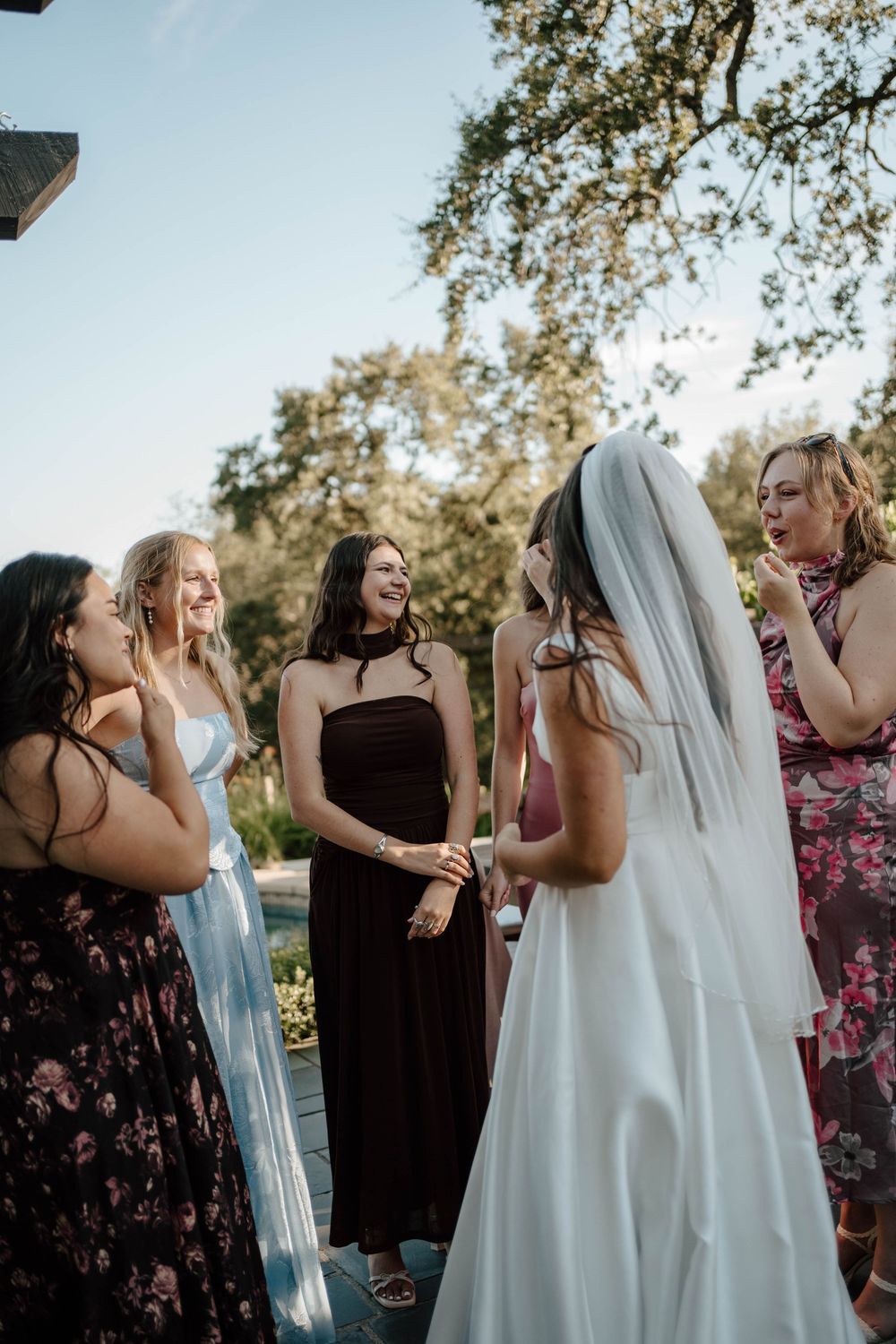 Bride in white gown and veil laughing with bridesmaids in colorful dresses outdoors at a wedding celebration.