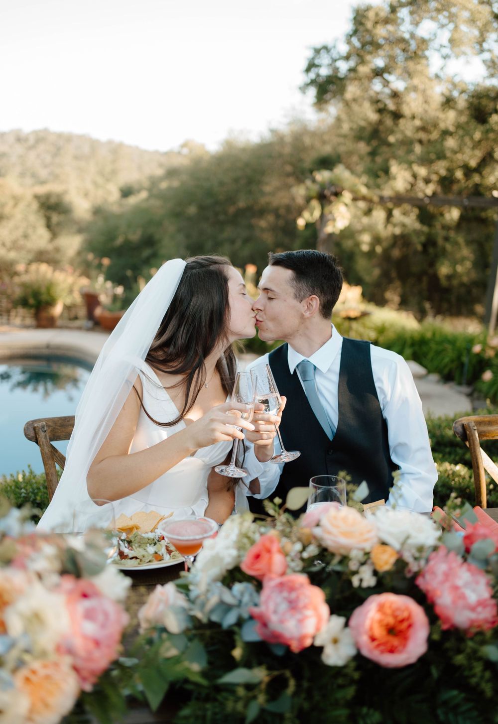 Bride and groom share a kiss while toasting champagne at an outdoor wedding reception table with colorful floral arrangements.