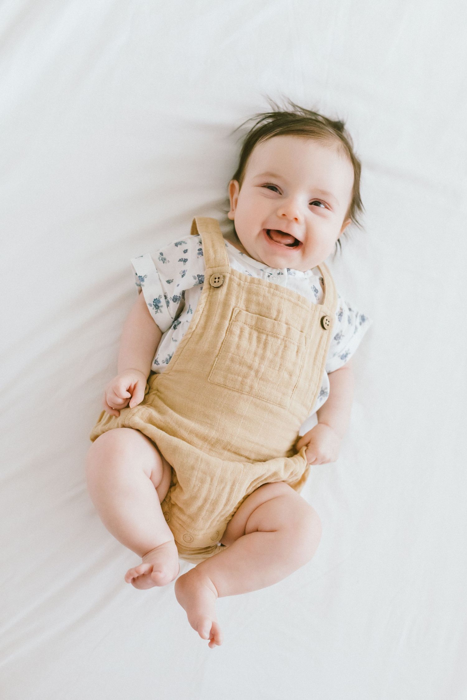 Smiling baby in tan overalls and floral shirt lying on white fabric, looking up with a joyful expression.