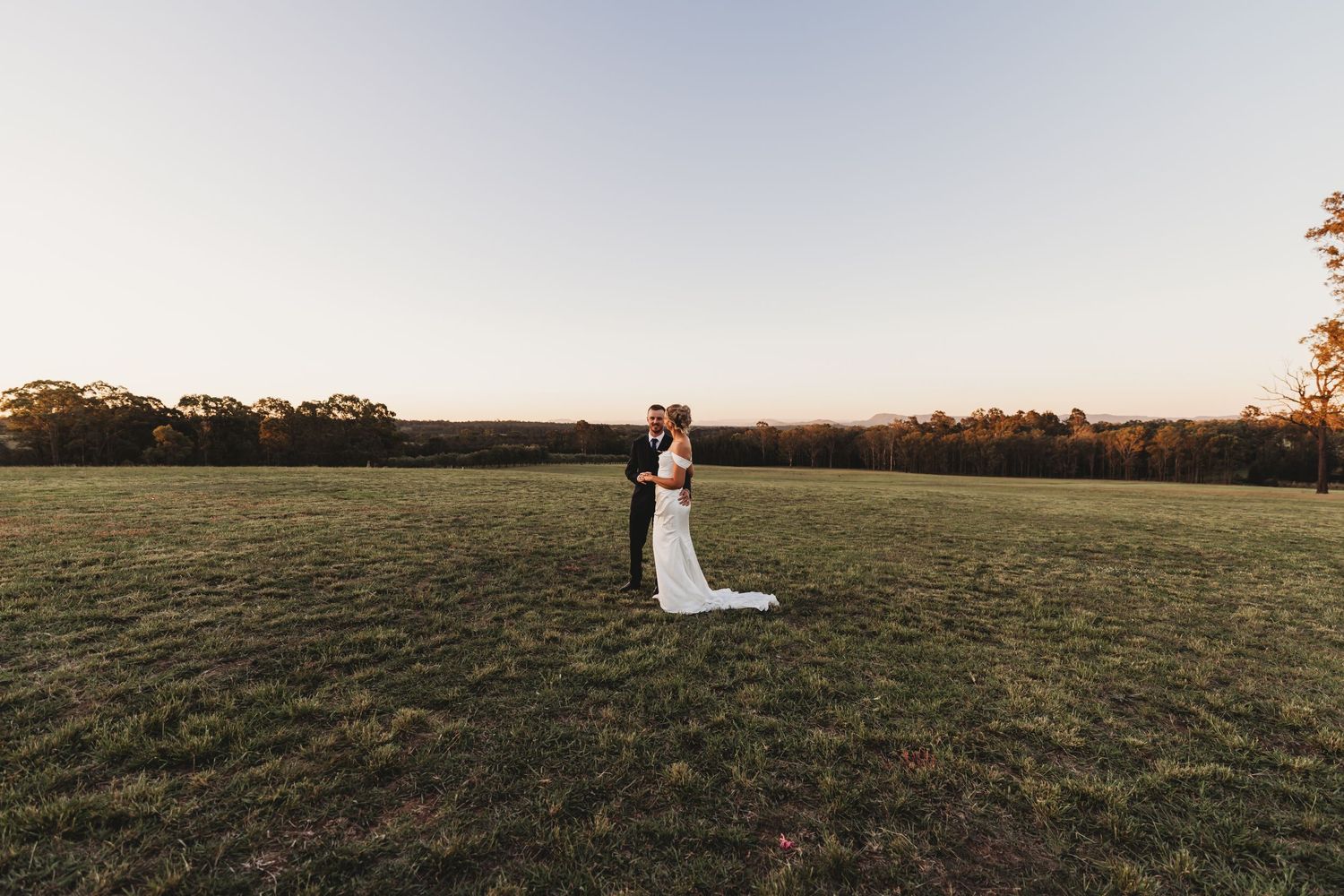 A bride and groom embrace in a vast green field at golden hour with trees and clear sky in the background.
