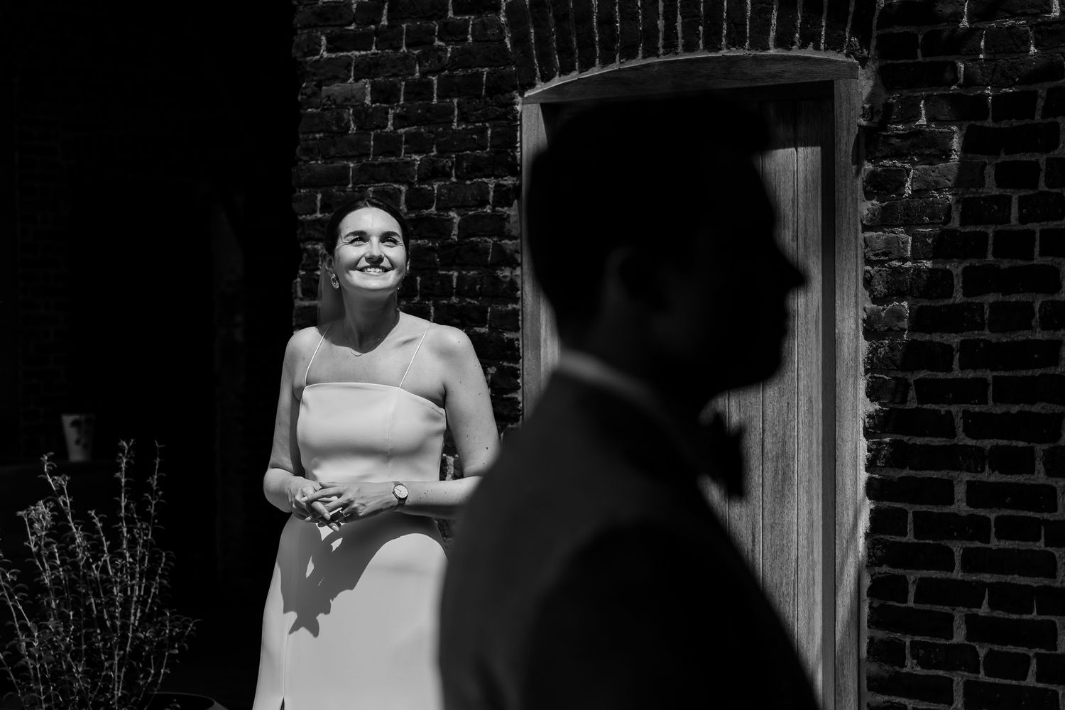 Black and white wedding photo of smiling bride in elegant gown looking at groom silhouette near brick wall.