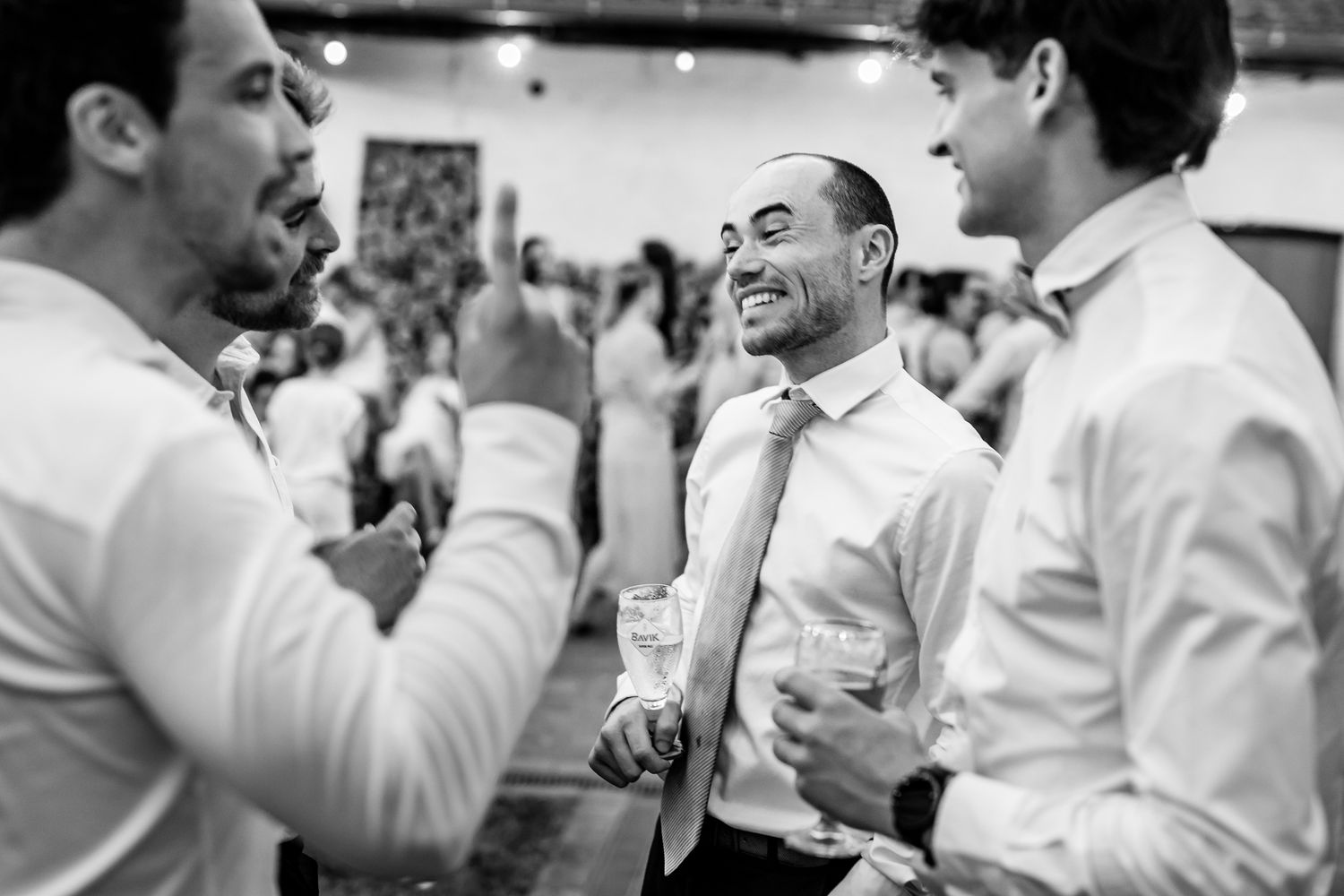 Men in white dress shirts laughing and socializing at a formal event, holding drinks in a candid black and white photo.
