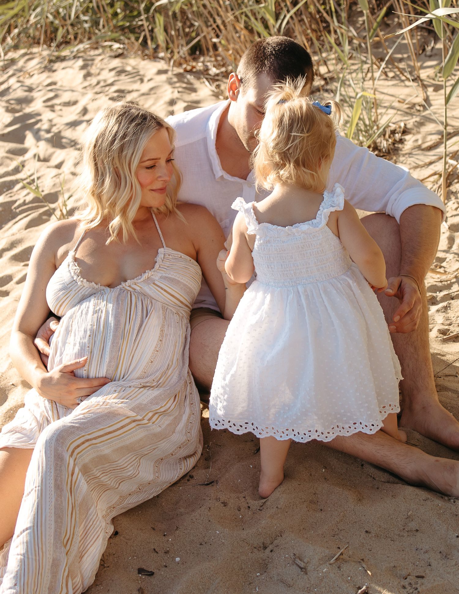 Pregnant woman in striped dress sits on beach with partner and toddler girl in white dress during golden hour.